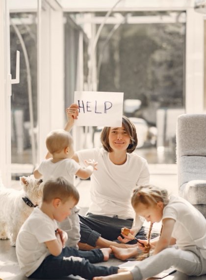A parent holding up a handwritten HELP sign while three children play around them