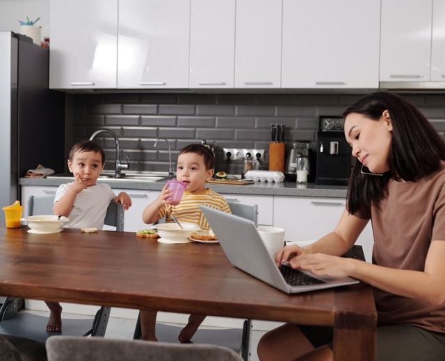 A parent working on a laptop while two children eat at the kitchen table